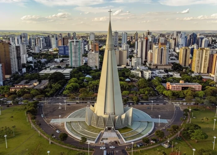 Vista aérea da Catedral de Maringá, uma imponente igreja em formato de cone feita de concreto. Ela está localizada no centro de uma grande praça circular com gramados e árvores, e é cercada por uma cidade densa com muitos prédios altos que se estendem até o horizonte. O céu está parcialmente nublado e a iluminação é de fim de tarde.