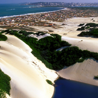 Vista aérea panorâmica de Natal, Rio Grande do Norte, destacando em primeiro plano uma lagoa azul cercada por dunas de areia branca e vegetação nativa, com a cidade e a costa litorânea estendendo-se ao fundo sob um céu ensolarado.