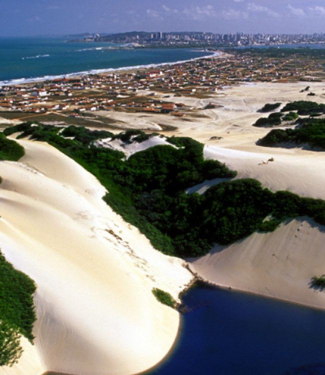 Vista aérea panorâmica de Natal, Rio Grande do Norte, destacando em primeiro plano uma lagoa azul cercada por dunas de areia branca e vegetação nativa, com a cidade e a costa litorânea estendendo-se ao fundo sob um céu ensolarado.
