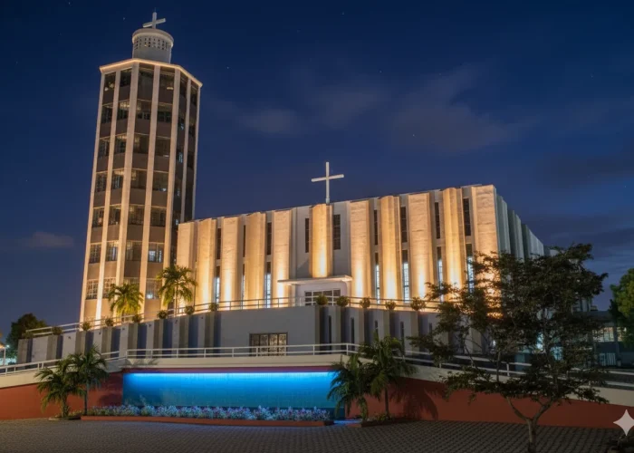 Fotografia noturna de uma grande igreja de arquitetura moderna. O edifício possui uma torre alta à esquerda e uma fachada com pilares verticais. Toda a estrutura é iluminada por luzes amarelas de baixo para cima, com uma luz azul intensa e contrastante na base, sob um céu escuro.
