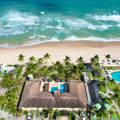Fotografia aérea vertical do Atlântico Sul Hotel em Porto de Galinhas, mostrando a fachada do hotel, sua área de piscina interna, carros estacionados na rua frontal, e uma ampla faixa de areia clara que se conecta ao mar azul com ondas cristalinas.