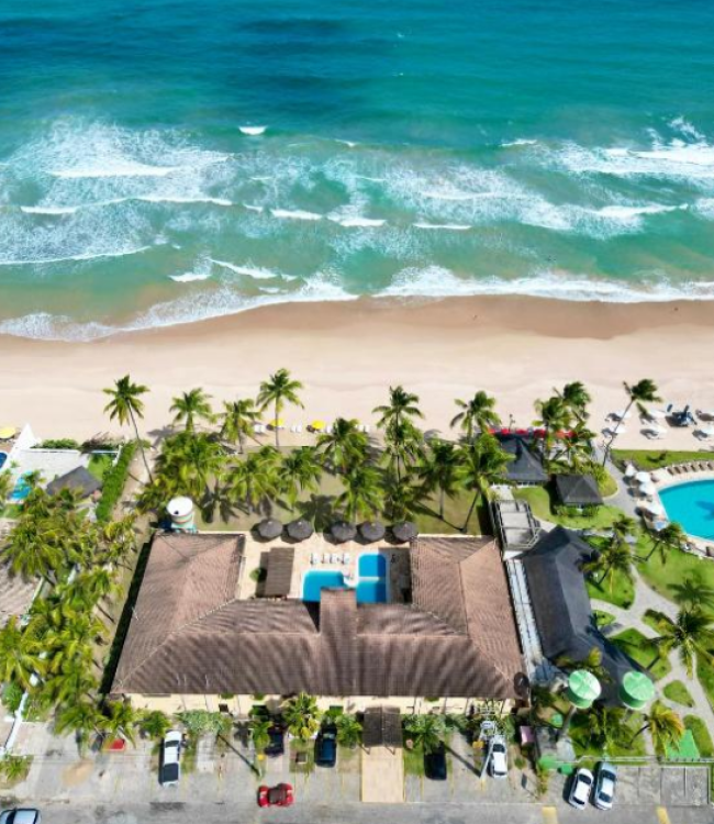 Fotografia aérea vertical do Atlântico Sul Hotel em Porto de Galinhas, mostrando a fachada do hotel, sua área de piscina interna, carros estacionados na rua frontal, e uma ampla faixa de areia clara que se conecta ao mar azul com ondas cristalinas.