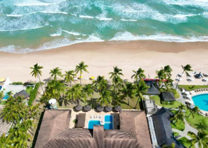 Fotografia aérea vertical do Atlântico Sul Hotel em Porto de Galinhas, mostrando a fachada do hotel, sua área de piscina interna, carros estacionados na rua frontal, e uma ampla faixa de areia clara que se conecta ao mar azul com ondas cristalinas.