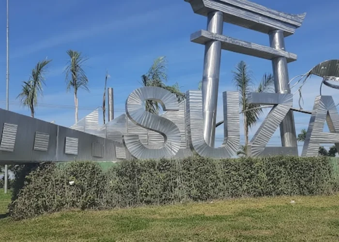 Uma imagem diurna de um grande monumento com a palavra "SUZANO" escrita em letras trateadas e tridimensionais, em um estilo moderno. As letras são altas e refletem a luz do sol, que brilha intensamente de um céu azul claro e sem nuvens. Elementos decorativos associados à cultura japonesa estão integrados na estrutura, incluindo um portão torii de metal prateado que se eleva sobre uma das letras e formas curvas que lembram as dobras de um origami ou um peixe koi estilizado em outras partes do monumento. Palmeiras esguias e algumas folhagens baixas pontuam o fundo, enquanto na base do monumento há um canteiro de arbustos baixos e densos, e na frente, um gramado verde e bem cuidado. A cena transmite uma sensação de brilho e orgulho cívico.