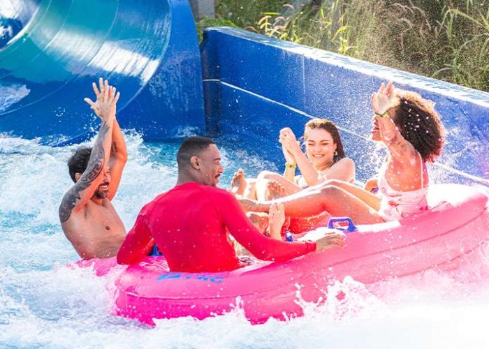 Quatro amigos em uma boia inflável rosa descendo um toboágua azul em um parque aquático hotpark. O grupo está sorrindo e se divertindo, com a água espirrando ao redor.
