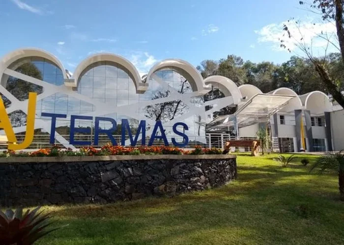 Fachada do AJ Termas Hotel em um dia ensolarado, mostrando sua arquitetura moderna com arcos e vidro. À frente, um letreiro azul com o nome "AJ TERMAS" sobre um canteiro de pedras escuras e flores. O hotel é cercado por um gramado verde e árvores.