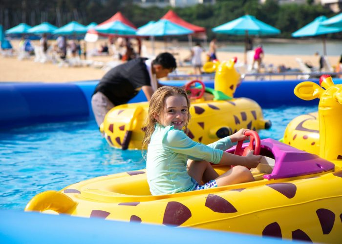 "Uma menina sorridente brinca em um barco inflável amarelo com formato de girafa em uma piscina infantil azul. Ao fundo, outras pessoas e brinquedos infláveis são visíveis, com cadeiras de praia e guarda-sóis coloridos em uma área de areia."